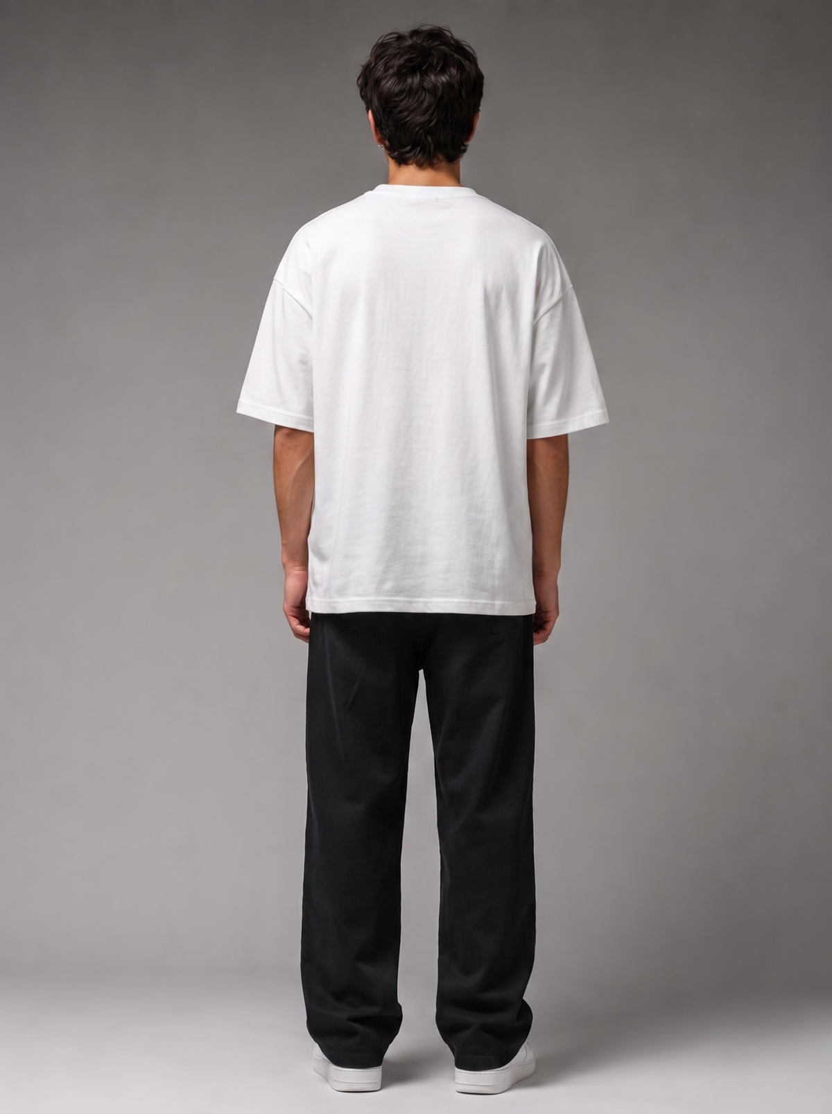 A man viewed from behind wearing an oversized white short-sleeve t-shirt and loose black pants, standing against a neutral gray studio background.