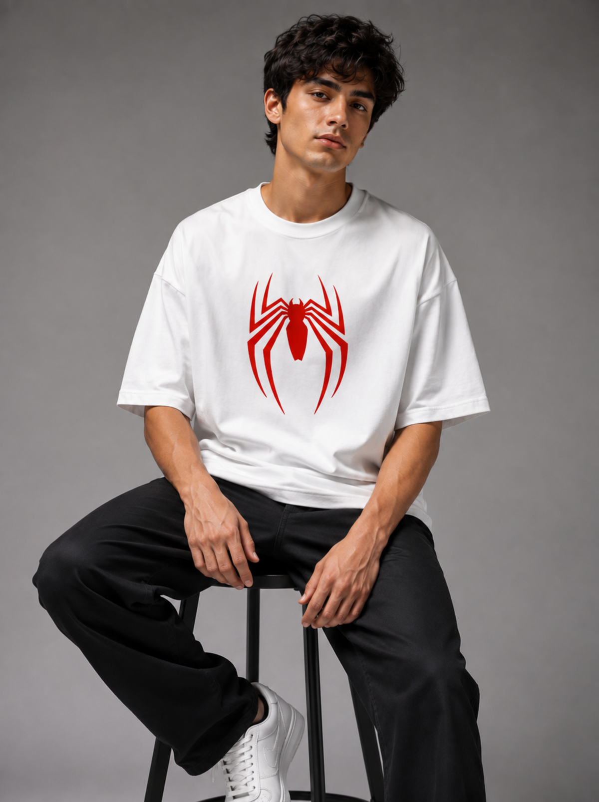 A man wearing a white crewneck t-shirt with a red spider logo on the chest, paired with black pants and white sneakers, sitting on a stool against a gray studio background.